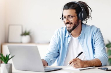 Mesafe öğrenme. Kablosuz kulaklıklı mutlu Hintli adam dizüstü bilgisayarı izliyor ve not alıyor, çevrimiçi eğitim ya da konferans veriyor, İçişleri Bakanlığı 'nda masa başında oturuyor, boş yer arıyor.
