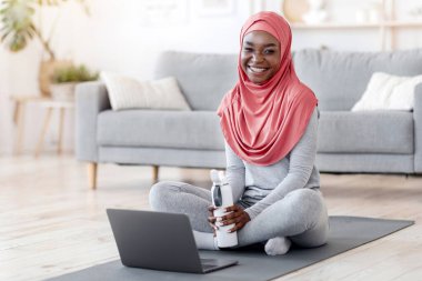 Çevrimiçi Yoga için hazır. tesettürlü, tesettürlü, tesettürlü, dizüstü bilgisayarın önünde oturan, video dersleriyle evde eğitim gören, kameraya gülümseyen siyah Müslüman kadın.