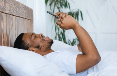 Neşeli Afro-Amerikalı adam telefonuyla interneti tarıyor cep telefonuyla evde, yatakta uzanıyor. Hafta sonu tatili, telefon konsepti için cep telefonu uygulaması. Yan Görünüm