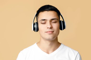 Boş zaman, meditasyon, rahatlama ve evde müzik eşliğinde dinlenme. Kulaklıklı sakin genç adam müzik listesini dinliyor, bej arka planda izole edilmiş, kapatın.