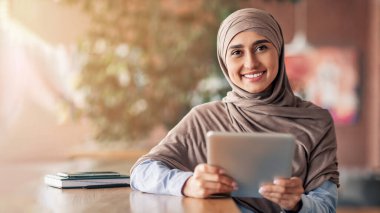 Dijital tabletle tesettürlü mutlu kız kameraya gülümsüyor, kafede tek başına oturuyor, boşluğu kopyalıyor.