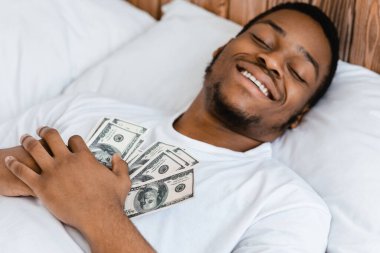 Mutlu Afrikalı Amerikalı adam elinde para tutarak uyur zenginlik hayali kurar ve evde yatarak zengin olur. Mali Bütçe, Mali Gelir ve Tasarruf Güvenliği Konsepti. Seçici Odaklanma