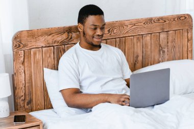 Serbest meslek hayatı. Afrikalı Amerikalı serbest çalışan adam dizüstü bilgisayarda çalışıyor internette internette internette evde yatakta oturuyor. Uzak Bilgisayar Görev Konsepti. Seçici Odaklanma