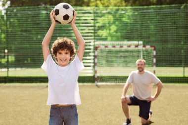 Aktif Gençlik Konsepti. Futbol topu tutan mutlu kıvırcık çocuk, çocuk parkında duruyor, babası arka planda oturuyor.