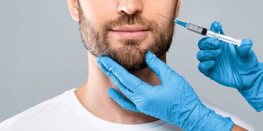 Tanımlanamayan sakallı adam güzellik salonunda dudak dolgusu yaptırıyor, gri stüdyo geçmişi var, kesilmiş. Kozmetik uzmanlarının elleri insan dudaklarına hyaluronik asit dolduruyor. Erkek kozmetik, estetik tıp