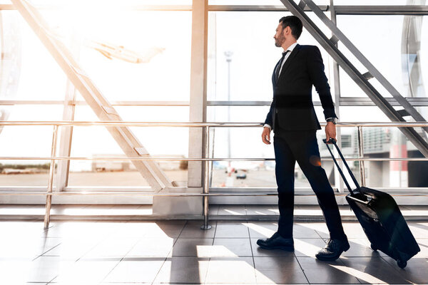 Красивый бизнесмен, прогуливающийся по терминалу аэропорта с багажом, смотрящий в окно на самолет, боковой вид, копировальное пространство. Предприниматель в черном костюме идет по современному аэропорту, солнечный свет