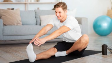 Spor, pilates ve jimnastik. Oturma odasında spor malzemeleriyle esneme hareketleri yapan bir adam.