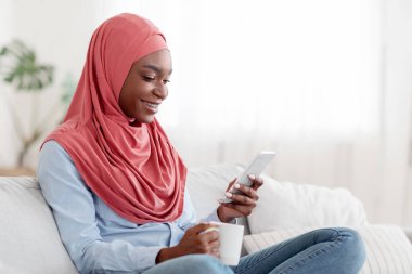 Gülümseyen siyah Müslüman kadın evde dinleniyor, akıllı telefon kullanıyor ve kahve içiyor oturma odasında kanepede dinlenirken, tesettüre girmiş dindar İslamcı kadın evdeki rahatlığın, boş alanın keyfini çıkarıyor.