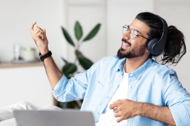 Be Yourself. Joyful weatern freelancer guy listening music in headphones and playing virtual guitar, relaxing at desk in home office, young indian man having fun and enjoying working remotely