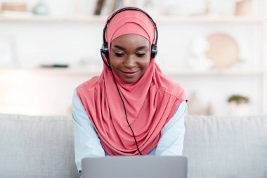 Evde dizüstü bilgisayar ve kulaklıkla çalışan, tesettürde oturan, oturma odasında oturan ve klavyede yazan Afrikalı Müslüman kadın, uzaktan kumandayla eğlenen,