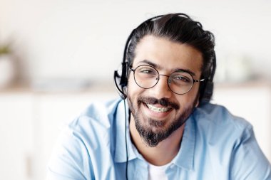 Merkez Operatörü 'nü ara. Pozitif Hintli adam müşteri hizmetleri temsilcisinin yakından çekilmiş portresi. Kulaklıklı yakışıklı batı adam yardım hattı yöneticisi kameraya bakıyor ve gülümsüyor, seçici odaklanıyor.