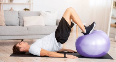 Karantina sırasında evde topla yapılan alıştırmalar. Spor giyim ve spor kıyafetleri giyen atletik adam oturma odasında karın kasları egzersizi yapıyor.