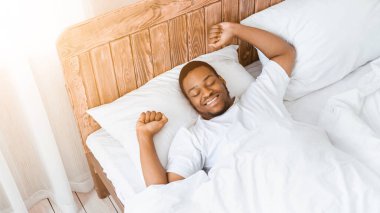 İyi uyuyan Afro-Amerikan bir adam uyanıyor ve elleri uzanıyor. Yatakta uzanmış, gözleri kapalı gülümsüyor. Sabah iyi bir uykudan sonra uyanmak, dinlenme ve dinlenme konsepti. Yüksek Açı, Panorama