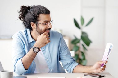 Genç Hintli erkek tasarımcı renk örneklerini tutuyor ve inceliyor, yaratıcı ofiste masa başında çalışıyor, iç tasarımı seçiyor, yeni proje için renk gama 'sını kontrol eden şık batılı adam, serbest alan