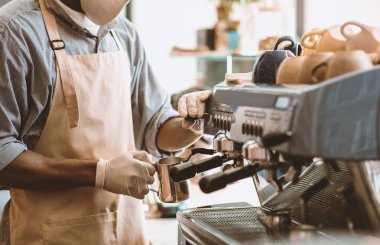 Kahve yapmak ve koronavirüs salgını sırasında yeni normaller. Milenyum Afrikalı Amerikalı, önlüklü, maskeli ve eldivenli barista sabahları kahve makinesinde sıcak içecek yapar.