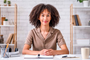 Covid-19 karantinası sırasında merkez ofis. Not defteri ve kulaklığı olan tatlı Afro-Amerikalı kız masada oturur ve oturma odasındaki kameraya bakar.