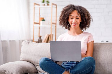 İnternette sörf yapmak ve evde iş aramak. Gülümseyen Afro-Amerikan genç kadın laptopta bilgi arıyor, oturma odasındaki koltukta oturuyor salgın sırasında, boş alan