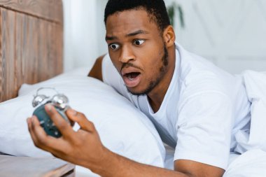 Uyuyakalmışım. Şok olmuş siyah bir adam, sabah işe geç saatte uyanıp, evde yatarken, bozuk alarma bakıyor. Uyanma Sorunu ve Gecikme, Kötü Sabah