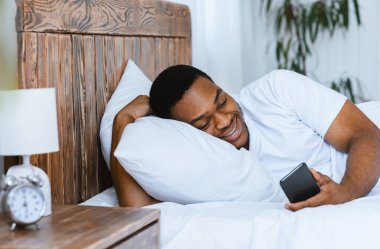 Mutlu Afrikalı adam cep telefonu kullanıyor çevrimiçi oyunlar oynuyor ve evde yatakta uzanmış akıllı telefonuyla interneti tarıyor. Hafta sonu Sabah Tatili, Mobil Uygulama Konsepti