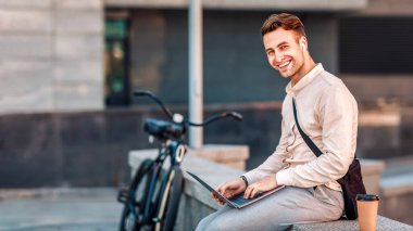 Açık hava şebekesi. Kablosuz kulaklıklı, kahveli mutlu adam şehirde dizüstü bilgisayarda çalışıyor ve kameraya bakıyor.