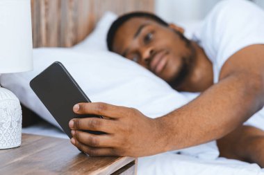 Siyah adam akıllı telefondan çalar saati kapatıyor. Sabahın köründe evde yatarak uyanıyor. Uyku eksikliği, dinlenme ve dinlenme konsepti. Seçici Odaklanma, Sığ Derinlik