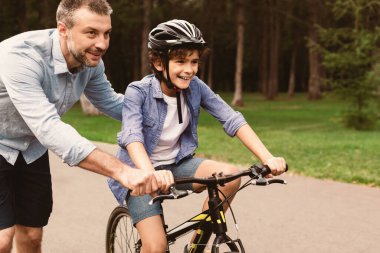 Destek konsepti. Gülümseyen bir babanın, pozitif oğluna bisiklet sürmeyi öğretirken, gidonu tutarken, hafta sonları dışarıda boş vakit geçirirken portresi. Çocuk öğrenirken eğleniyor.
