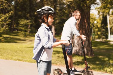Paylaşım Servisi. Şirin kıvırcık çocuk babasıyla elektrikli scooter kullanıyor. Doğada, kenarda, seçici bir odak noktasında.