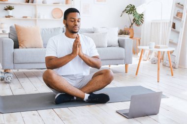 Afro-Amerikalı adam yoga yapıyor laptopun başında oturuyor ve evde Lotus pozisyonunda meditasyon yapıyor. Çevrimiçi Meditasyon ve Rahatlama, Sakin Kalın Konsepti. Seçici Odaklanma