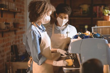 Barista sosyalleşme ve covid-19 salgınında çalışıyor. Milenyum Afrikalı Amerikalı kadın ve önlüklü ve koruyucu maskeli adam kafenin içinde, boş bir yerde, barın arkasında kahve makinesi kullanıyorlar.