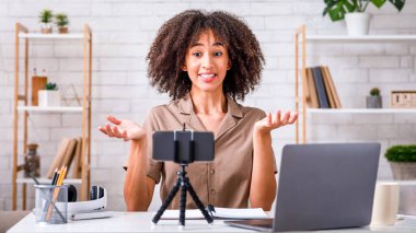 Bilgi, ders, hatalar ve online sorular. Şaşırmış Afrikalı Amerikalı kadın kollarını yana açıyor ve akıllı telefon web kamerasından izliyor. Oturma odasında dizüstü bilgisayarla oturuyor.