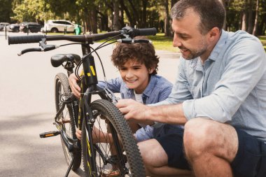 Kamu parkında tel kesiciyle bisiklet tamir eden neşeli baba ve oğlunun yakın plan portresi.