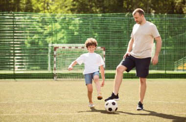 Baba ve oğlu topla futbol oynuyorlar. Güneşli bir günde şehir parkında yeşil alan, kopyala