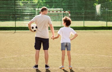 Yetişkin bir adam ve kıvırcık bir çocuğun, futbol topuyla spor sahasında el ele tutuşarak boy boy portresi.