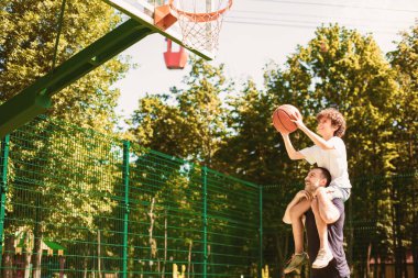 Destek. Olgun bir adam, küçük bir çocuğu omuzlarında tutuyor. Spor sahasında basket atmasına yardım ediyor.