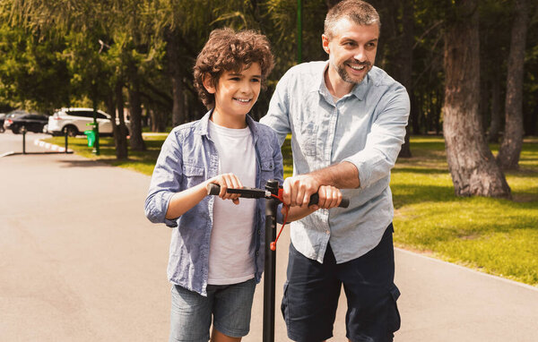 Концепция помощи. Портрет отца, который учит своего мальчика кататься на велосипеде в общественном парке, толкает электронный скутер, свободное место