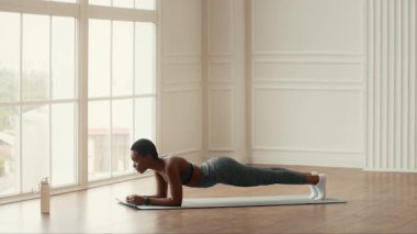 Kararlı bir Afrikalı Amerikalı kadın kapalı alanda bir tahta egzersizi yapar, atletizmini ve sağlıklı bir yaşam tarzına olan bağlılığını gösterir. Güneş ışığı odayı dolduruyor, çalışma ortamını geliştiriyor..