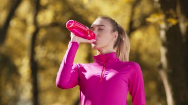 Atletik sarışın kadın, halka açık parkta çalışırken şişeden su içiyor.