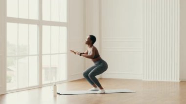 Bin yıllık Afrikalı Amerikalı bir kadın evde egzersiz yapıyor, yoga minderinde çömeliyor. Etrafı büyük pencerelerden gelen doğal ışıklarla çevriliyken spor rutinine odaklanıyor..