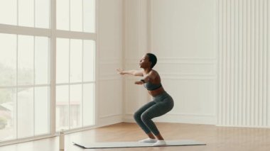 Bin yıllık Afrikalı Amerikalı bir kadın evindeki yoga minderinde çömeliyor. İyi ışıklandırılmış, geniş pencereli bir odada, sağlıklı bir yaşam tarzını teşvik etmeye odaklanıyor..