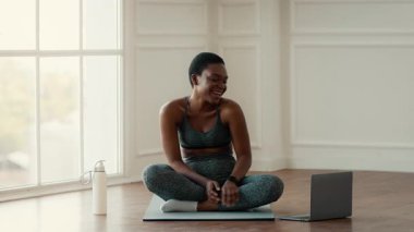 Milenyum Afrikalı Amerikalı bir kadın güneşli bir odada dizüstü bilgisayarını çalıştırırken sırıtarak bacak bacak üstüne atarak oturuyor..