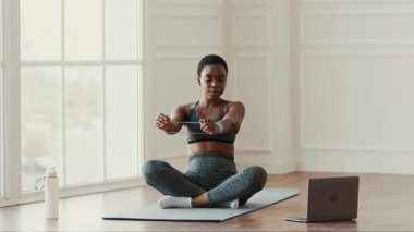 Bir kadın evde online antrenman yaparken bacak bacak üstüne atarak oturuyor. Sağlıklı bir yaşam tarzı sürdürmeye odaklanmış bir direniş bandı kullanıyor. Oda doğal ışıkla aydınlanmış..