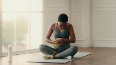 Evde spor yapan bir kadın mindere oturup bel ölçüsünü alıyor. Sağlıklı bir yaşam tarzını benimsiyor. Parlak ve davetkar bir ortamda ilerlemesini yansıtıyor..