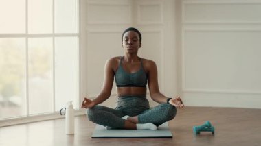 Bin yıllık Afro-Amerikan bir kadın bir paspasın üzerinde bacak bacak üstüne atmış, iyi aydınlatılmış bir odada meditasyon yapıyor. Ağırlıklar ve su şişesi yakınlarda, sağlıklı bir yaşam tarzına olan bağlılığını vurguluyor..