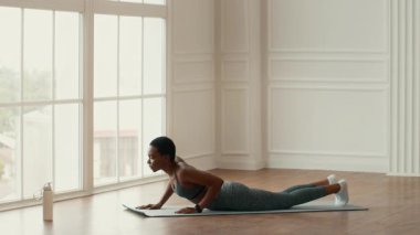 Afrikalı bir Amerikalı kadın içeride spor yapıyor, minder üzerinde yoga pozu veriyor. Doğal ışık geniş pencerelerden akarak atletik egzersizi için sakin bir atmosfer yaratır..