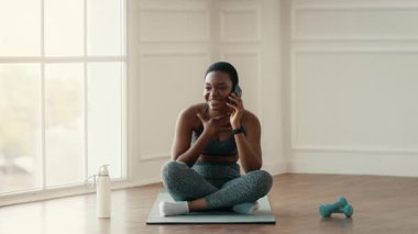 Parlak bir kapalı alanda, genç bir Afrikalı Amerikalı kadın spor minderinde bacak bacak üstüne atarak oturuyor, telefonda konuşurken gülümsüyor. Sağlıklı bir yaşam tarzını ve spor programını benimsiyor..