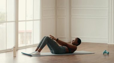 Genç bir Afrikalı Amerikalı kadın evdeki spor minderinde karın egzersizi yapıyor. Konsantrasyonunu koruyor, kendini spor salonuna adamış gibi görünüyor. Parlak bir mekanda..