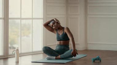 Genç bir Afrikalı Amerikalı kadın, kapalı alanda esneme egzersizleri yapıyor. Yoga minderinin üzerinde otururken sağlıklı bir yaşam tarzına odaklanır..
