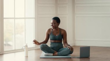 Bir kadın hasırda bağdaş kurup evde antrenman yapmaya hazırlanıyor. Yakınlarda bir dizüstü bilgisayar ve su şişesi olan, ışık dolu bir mekanla çevrili, enerjik ve mutlu görünüyor..