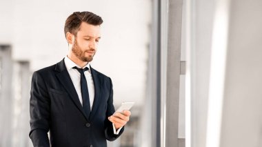 Yakışıklı sakallı iş adamı uçuş beklerken e-postayı kontrol ediyor, havaalanının penceresinin yanında duruyor, kopyalama alanı. Akıllı telefon kullanarak şık takım elbiseli orta yaşlı girişimci