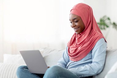 İnternet alışverişi. Başörtülü genç Müslüman kadın evde dizüstü bilgisayarla bir şeyler satın alıyor, web sitesinden eşyalar alıyor, oturma odasında kucağında bilgisayarla oturuyor, boşluğu kopyalıyor.